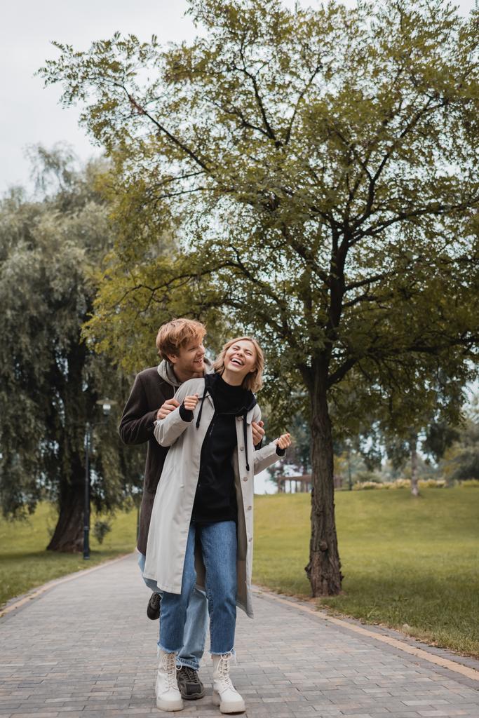 hombre feliz en abrigo abrazando alegre joven mujer con los ojos cerrados riendo en el parque  - Foto, imagen