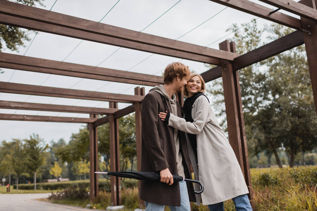 Улыбающаяся молодая пара в осенних пальто, обнимающаяся под множественной аркой в парке - Фото, изображение