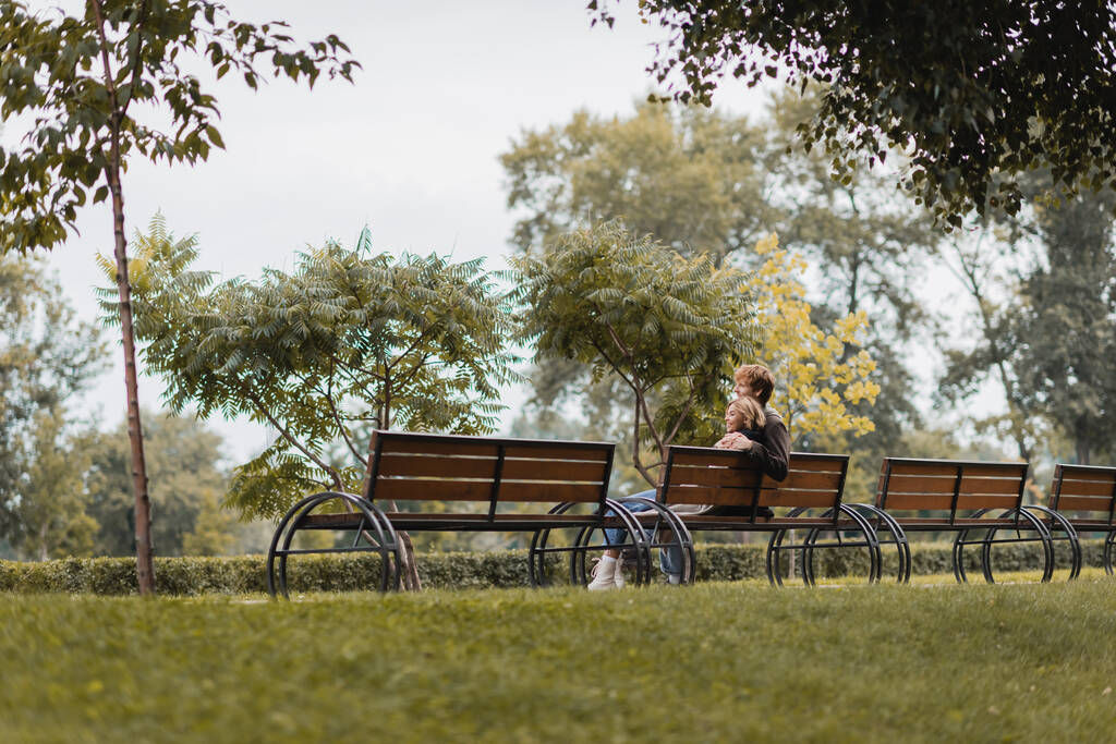 vrolijk jong man en vrouw knuffelen en zitten op houten bank in groen park - Foto, afbeelding