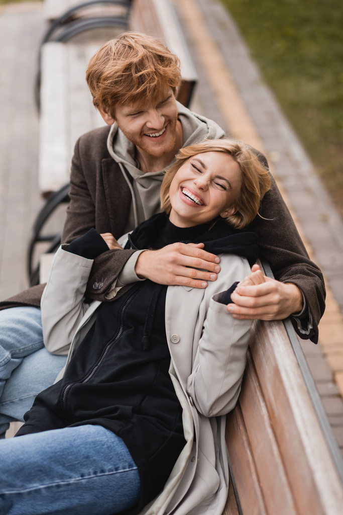 vista ad alto angolo di allegro giovane uomo che abbraccia donna sorridente e seduta su una panchina di legno nel parco - Foto, immagini