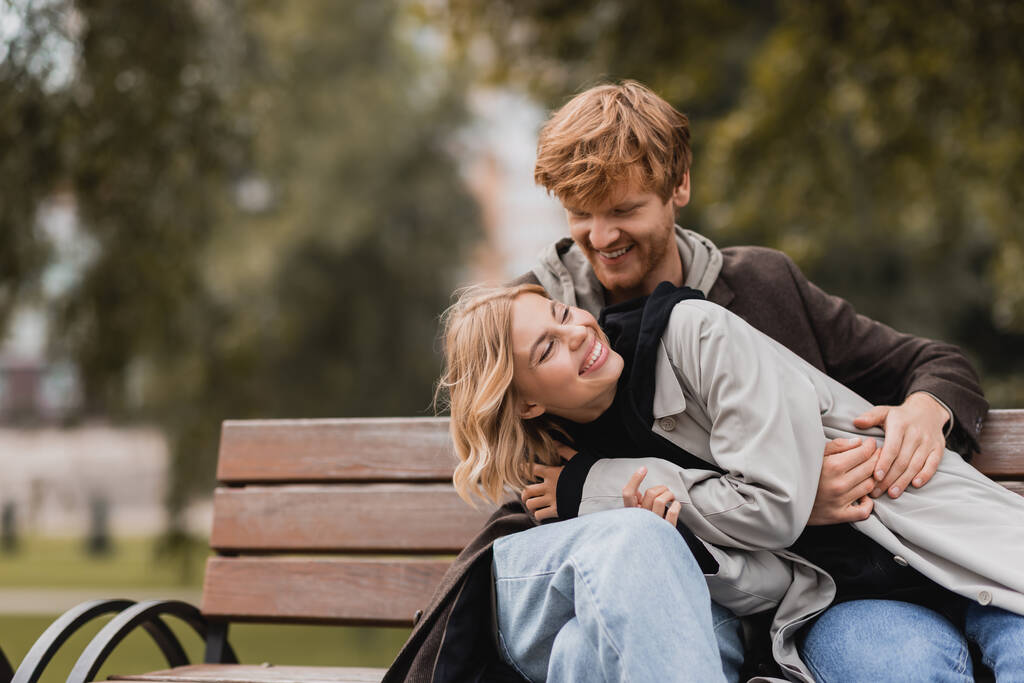 rothaariger Mann lächelt, während er fröhliche Freundin umarmt, während er auf Bank sitzt - Foto, Bild