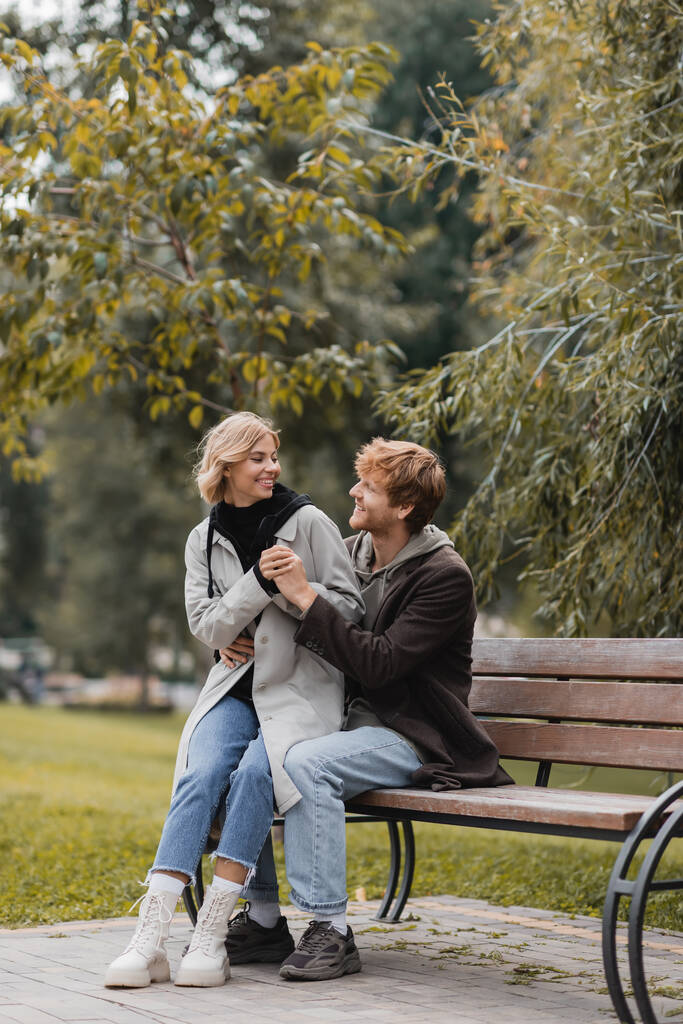 rousse homme souriant tout en embrassant petite amie positive tout en étant assis sur le banc - Photo, image