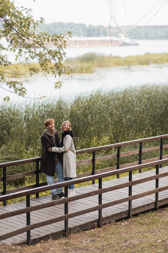 volledige lengte van roodharige man en gelukkige vrouw in jas glimlachen terwijl knuffelen op brug in de buurt van meer in park  - Foto, afbeelding