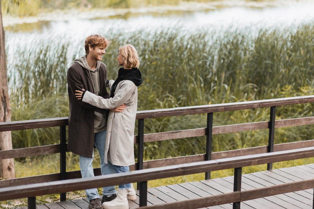 pełna długość ruda mężczyzna i blondynka w płaszcz uśmiecha się podczas przytulania na moście w pobliżu jeziora w parku  - Zdjęcie, obraz