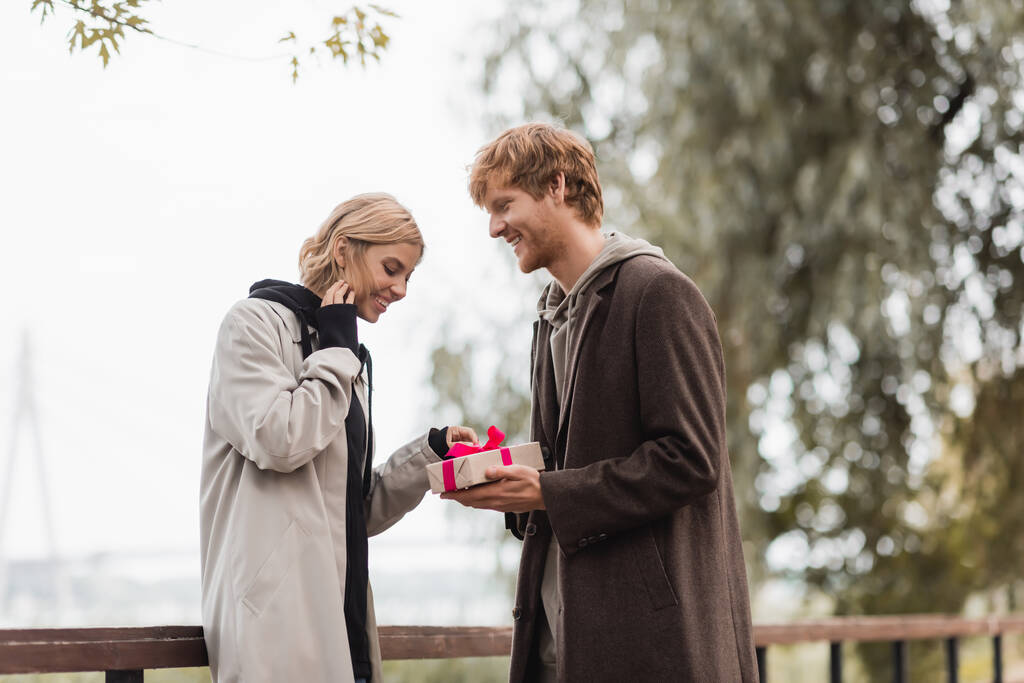 счастливый рыжий мужчина держит завернутый подарок около веселой девушки в парке - Фото, изображение