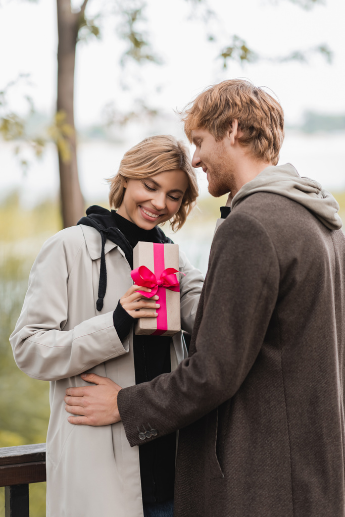 mujer feliz sosteniendo envuelto caja de regalo con cinta rosa cerca de novio alegre en el parque - Foto, Imagen