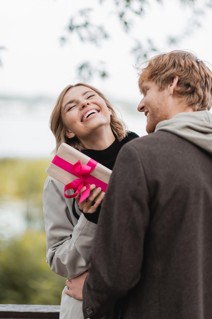 femme heureuse riant tout en tenant boîte cadeau enveloppé près copain gai dans le parc - Photo, image