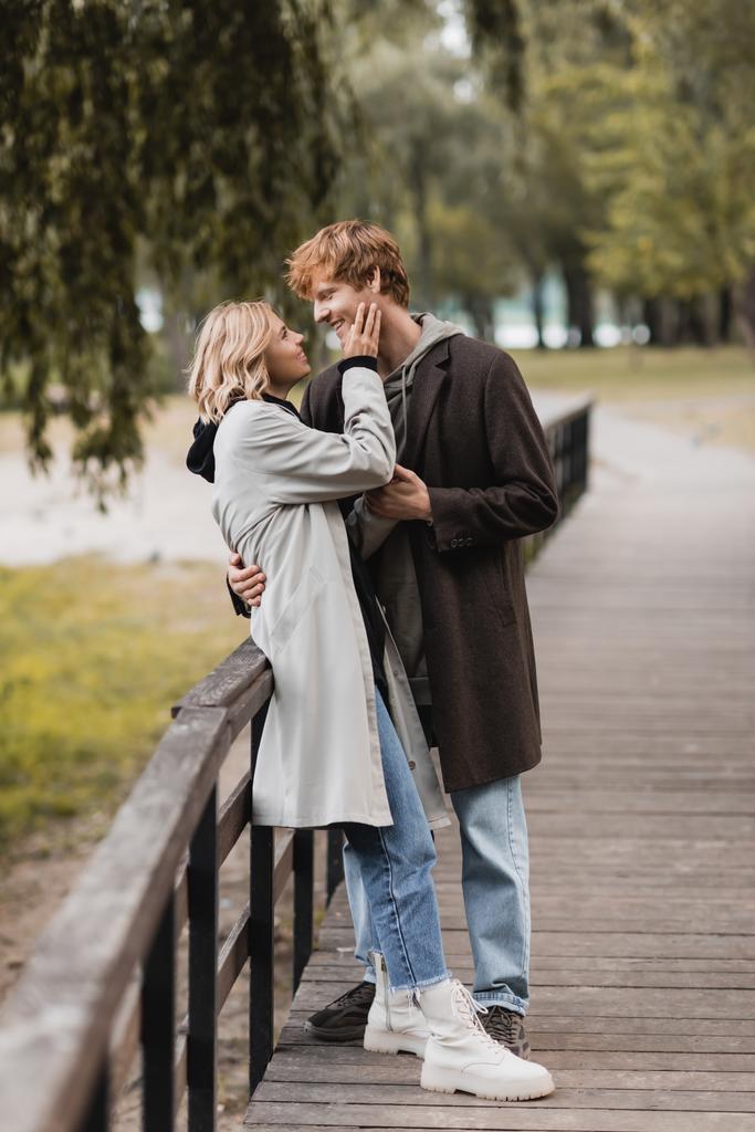 volle Länge des rothaarigen Mannes und der blonden Frau im Mantel, die sich beim Date im Park umarmen  - Foto, Bild