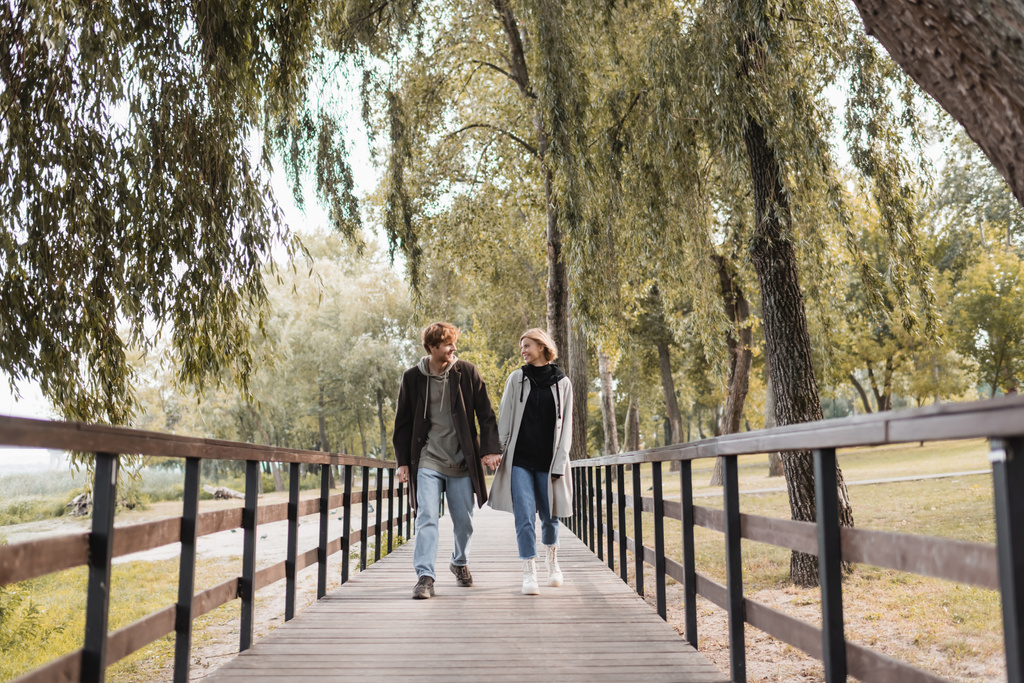 volledige lengte van roodharige man en blonde vrouw in jassen glimlachen terwijl het vasthouden van handen en lopen op de brug  - Foto, afbeelding