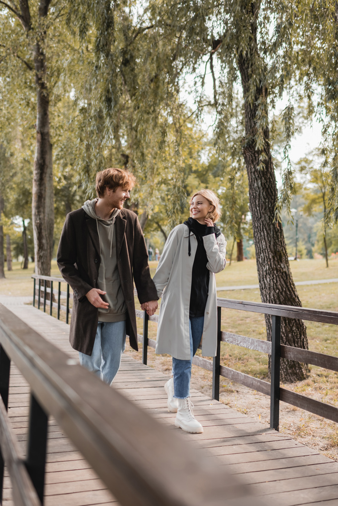 pełna długość szczęśliwej pary w płaszczach trzymających się za ręce i spacerujących po moście w parku - Zdjęcie, obraz