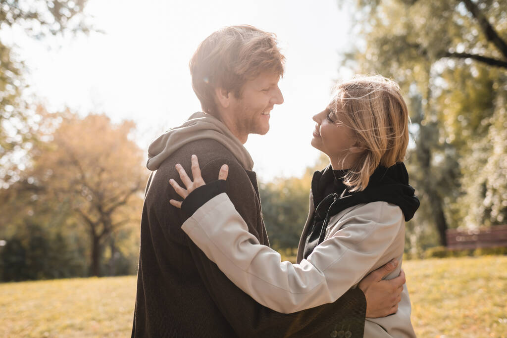 πλαϊνή άποψη του νεαρού και ευτυχισμένου ζευγαριού χαμογελώντας ενώ αγκαλιάζει στο φθινοπωρινό πάρκο  - Φωτογραφία, εικόνα