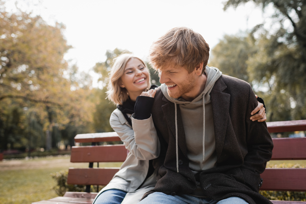 gelukkig en blond vrouw knuffelen roodharige vriend terwijl zitten op bank in park - Foto, afbeelding
