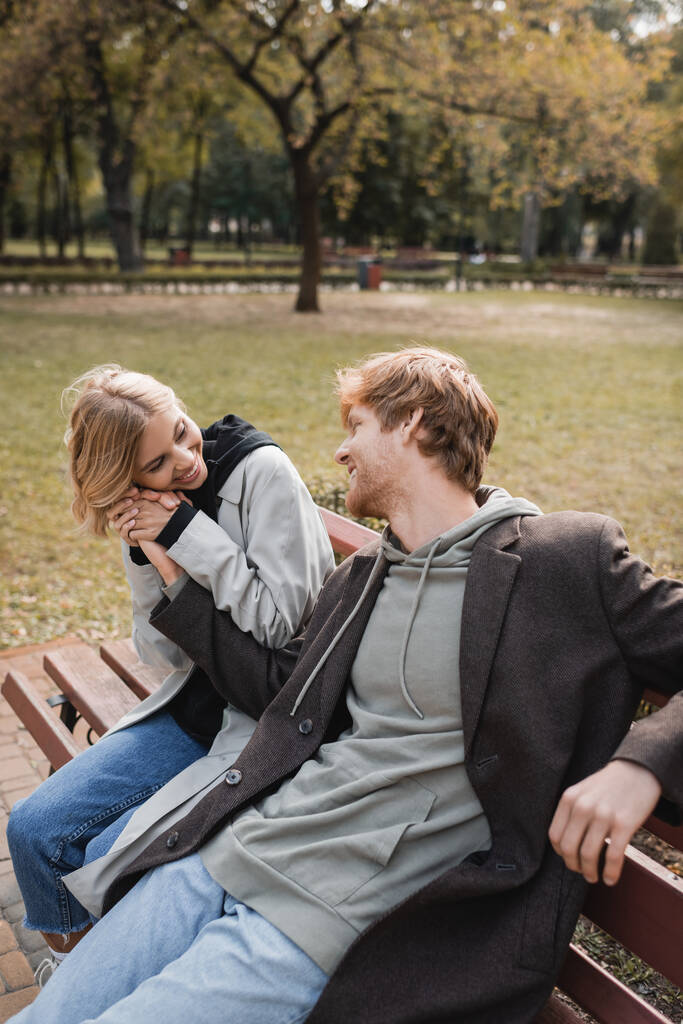 Parkta bankta otururken kızıl saçlı erkek arkadaşının kollarına sarılan neşeli ve sarışın kadın. - Fotoğraf, Görsel