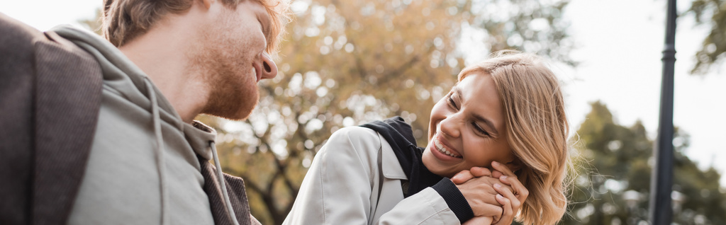 vrolijke en blonde vrouw houden hand van roodharige vriend terwijl glimlachen in het park, banner - Foto, afbeelding