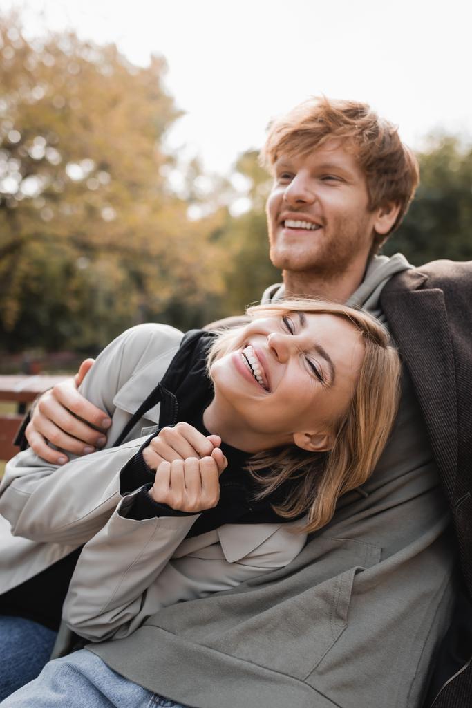 Rotschopf-Mann umarmt glückliche blonde Frau, während er im herbstlichen Park lächelt  - Foto, Bild
