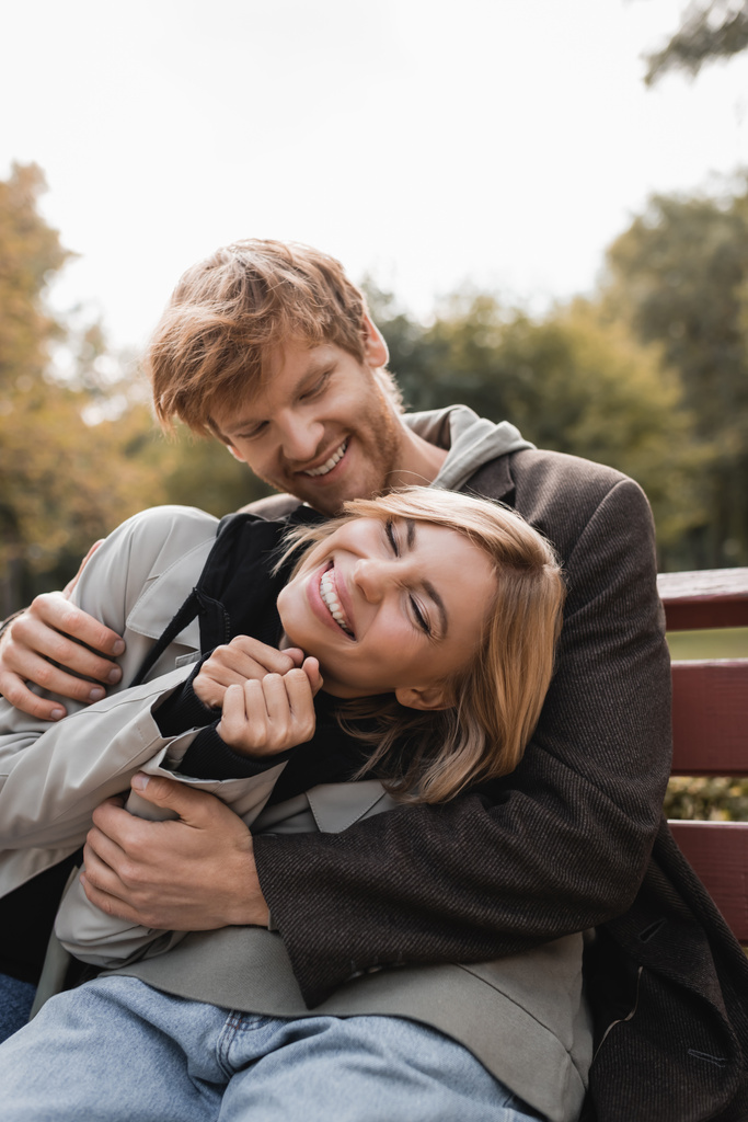 rothaariger junger Mann umarmt freudige blonde Frau, während er auf Bank im herbstlichen Park sitzt  - Foto, Bild