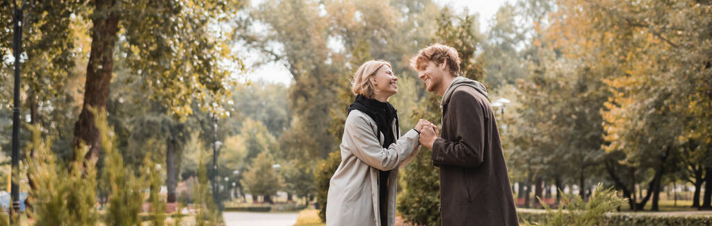 vista lateral de casal alegre e jovem em casacos de mãos dadas enquanto em pé em torno de plantas no parque, banner - Foto, Imagem