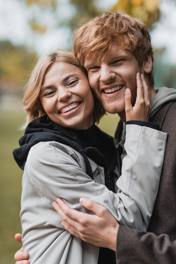 portrét šťastného páru v kabátech při pohledu na fotoaparát při objímání během rande - Fotografie, Obrázek