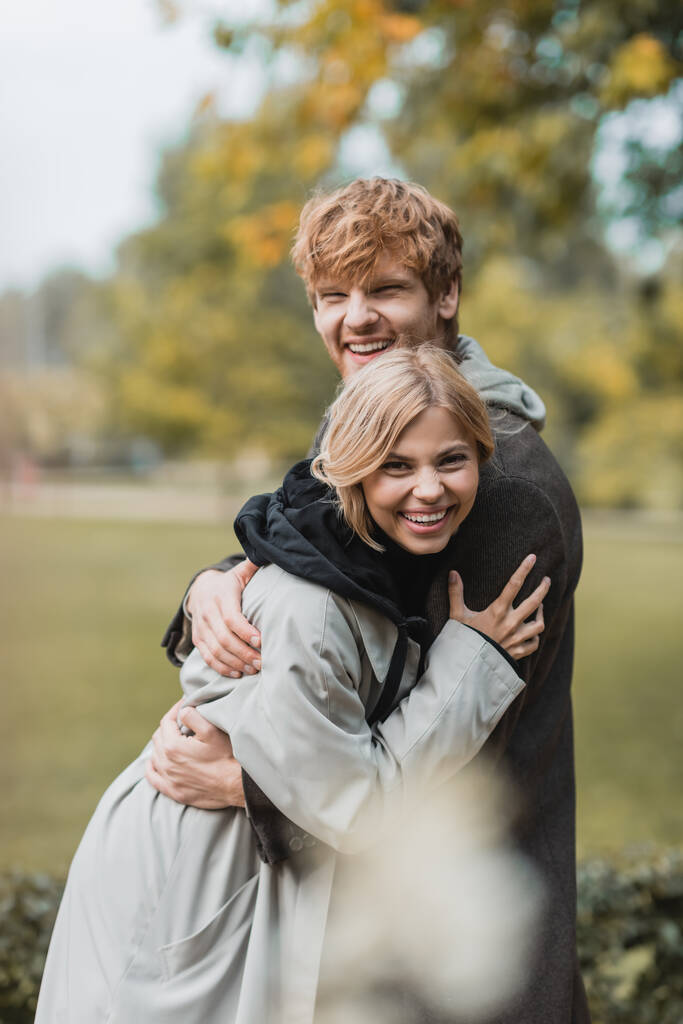 весёлый молодой человек и женщина в осенних пальто обнимают друг друга и улыбаются в парке - Фото, изображение