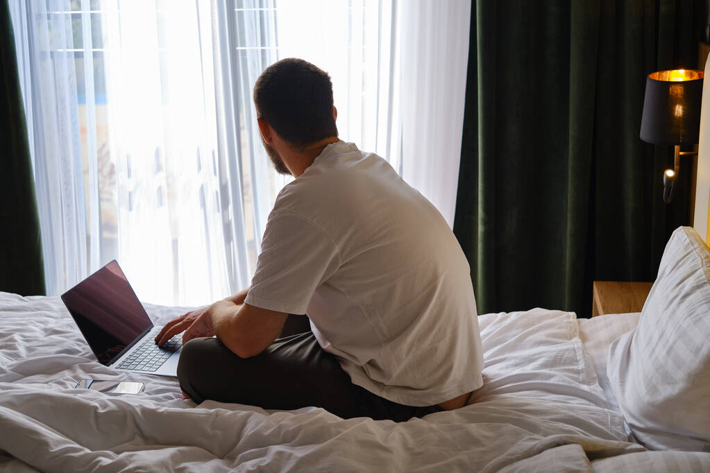 Muž pracuje s laptopem a smartphonem doma v posteli nebo v hotelech. Vzdálená práce. Kancelář doma. Muž v ložnici píše na notebook, dálkové studium pro studenty, surfování na internetu - Fotografie, Obrázek