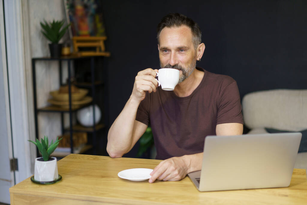Freelancer brodaty człowiek w domu pije kawę trzymając kubek w ręku siedzi przy biurku z komputerem PC - Zdjęcie, obraz