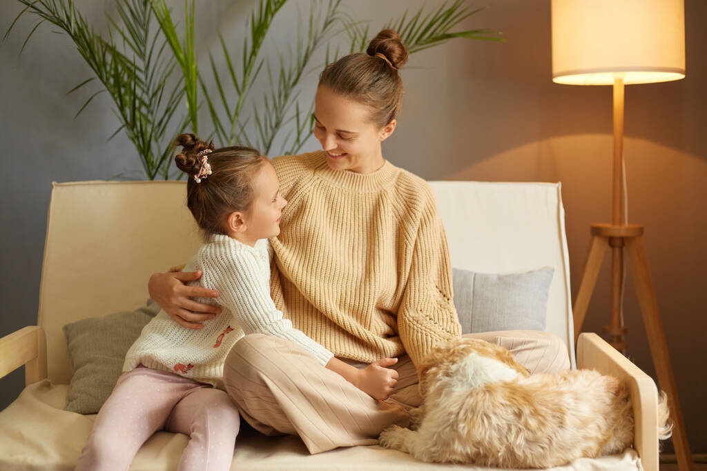 Horizontal tiro de feliz positivo otimista mãe e filhinha sentados no sofá e abraçando uns aos outros, aproveitando o tempo juntos e brincando com seu cão de Pequim. - Foto, Imagem