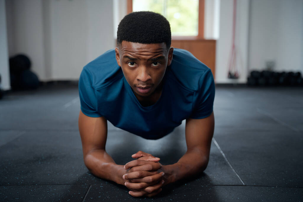 Сосредоточенный молодой черный мужчина в спортивной одежде делает упражнения на доске с руками, сжатыми в спортзале - Фото, изображение