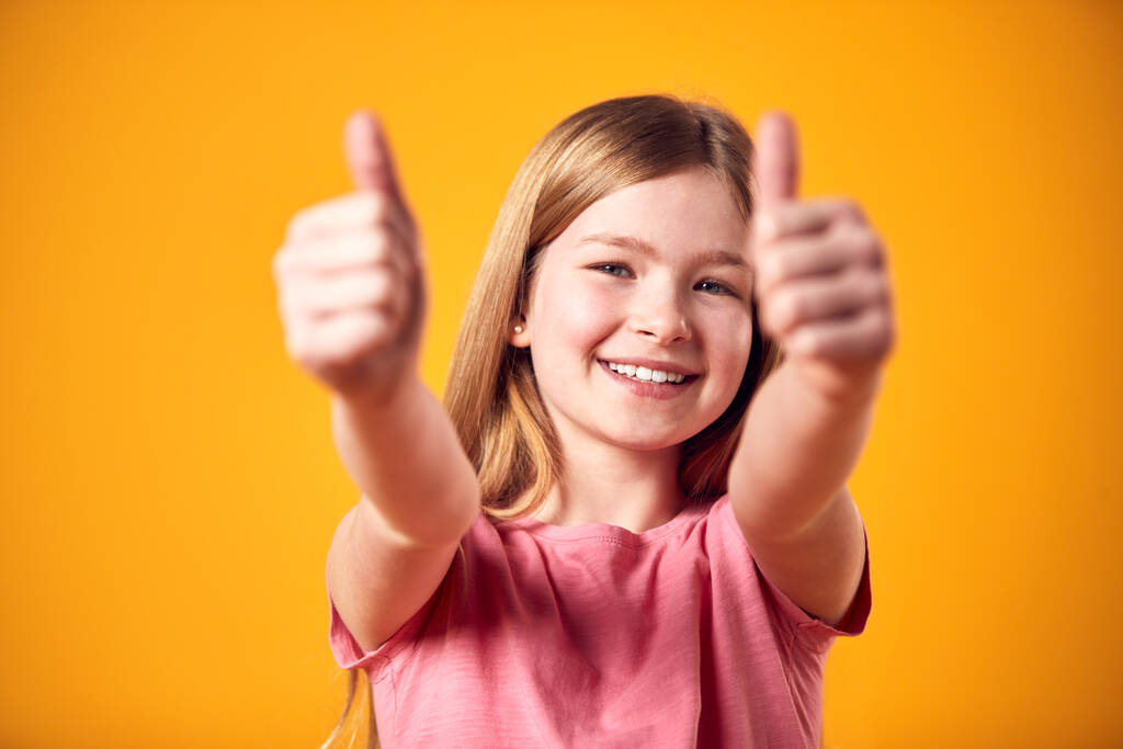 Studioporträt eines lächelnden jungen Mädchens, das vor gelbem Hintergrund Daumen hoch macht - Foto, Bild