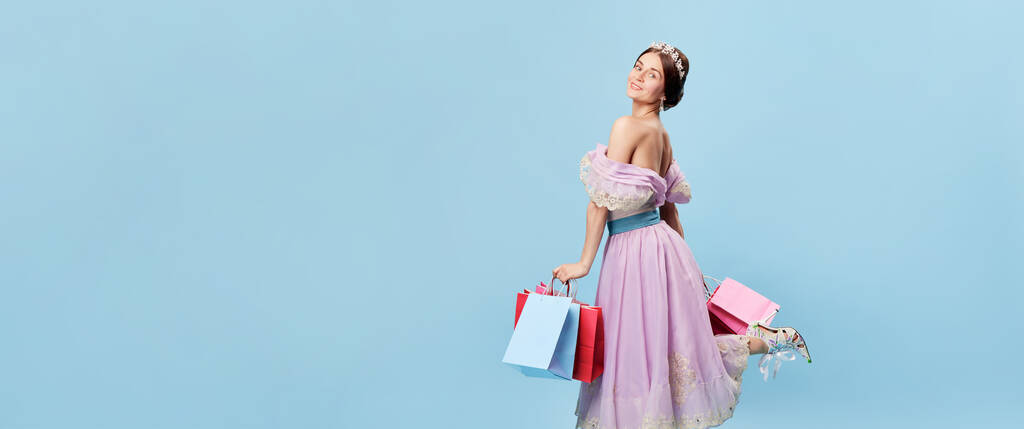 Flyer met charmante jonge vrouw in beeld van jonge koningin of prinses met kleurrijke boodschappentassen geïsoleerd op blauwe achtergrond. Menselijke emoties, schoonheid, zwarte vrijdag, kunst, verkoop, creativiteit. - Foto, afbeelding