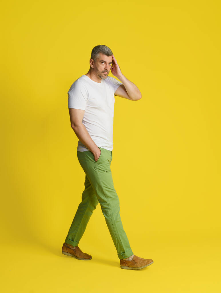 黄色の背景に隔離された白いTシャツと緑のジーンズを身に着けて手で中世のハンサムな灰色の髪の男のタッチの顔を歩く。フィット感、筋肉質の中年男性. - 写真・画像