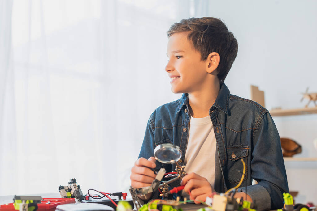 Sonriente niño preadolescente sosteniendo lupa cerca del modelo de robótica en casa  - Foto, imagen