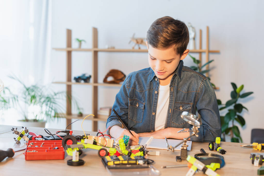Подросток пишет на тетрадке рядом с инструментами и роботизированной моделью дома  - Фото, изображение