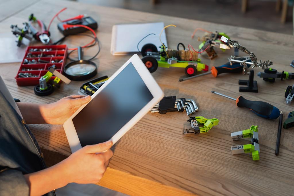 Teilansicht des Kindes, das ein digitales Tablet mit leerem Bildschirm in der Nähe von Werkzeugen und mechanischen Details auf dem Tisch hält - Foto, Bild