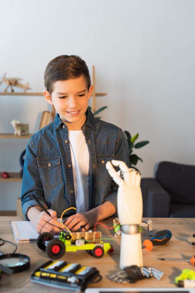 Glücklicher Junge betrachtet Roboterhand in der Nähe von Werkzeugen und mechanischen Teilen auf dem heimischen Tisch - Foto, Bild