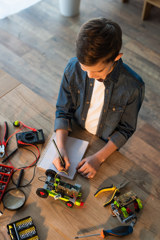 top view of boy writing in notebook near tools and details of robotics model on table - Photo, Image
