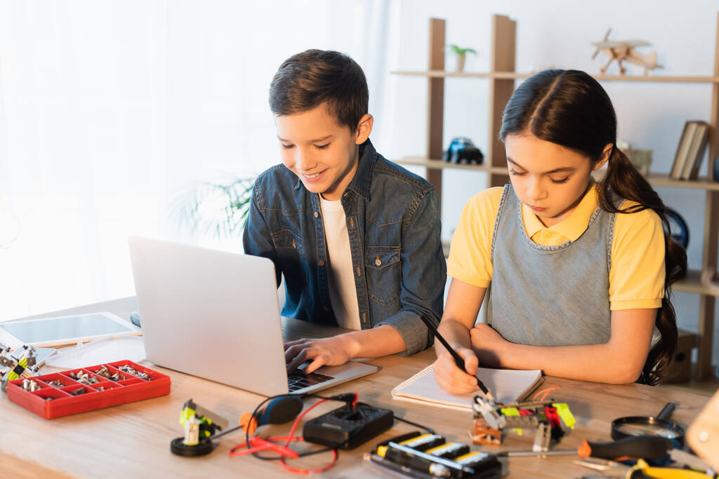 boldog fiú használ laptop, míg a lány írás notebook közelében részleteket robotika modell - Fotó, kép