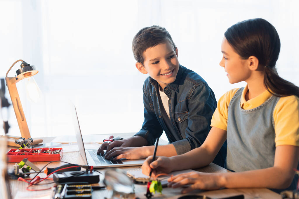 uśmiechnięty chłopiec korzystający z laptopa patrząc na dziewczynę piszącą w pobliżu rozmytych części modelu robotyki - Zdjęcie, obraz