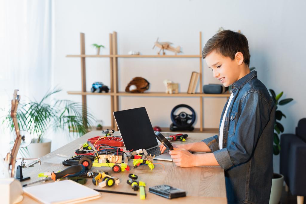 vista lateral del niño con lupa cerca de los detalles del modelo de robótica y el ordenador portátil con pantalla en blanco - Foto, Imagen