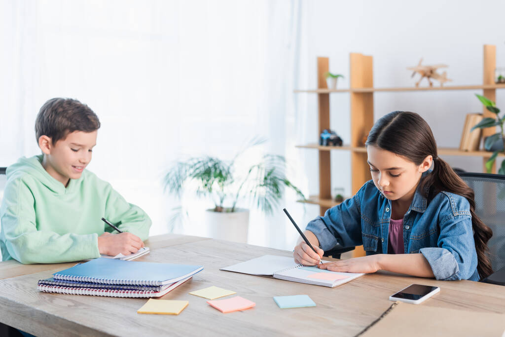χαμογελαστό αγόρι και συγκεντρωμένο κορίτσι γράφοντας σε σημειωματάρια κοντά σε άδειες αυτοκόλλητες σημειώσεις - Φωτογραφία, εικόνα