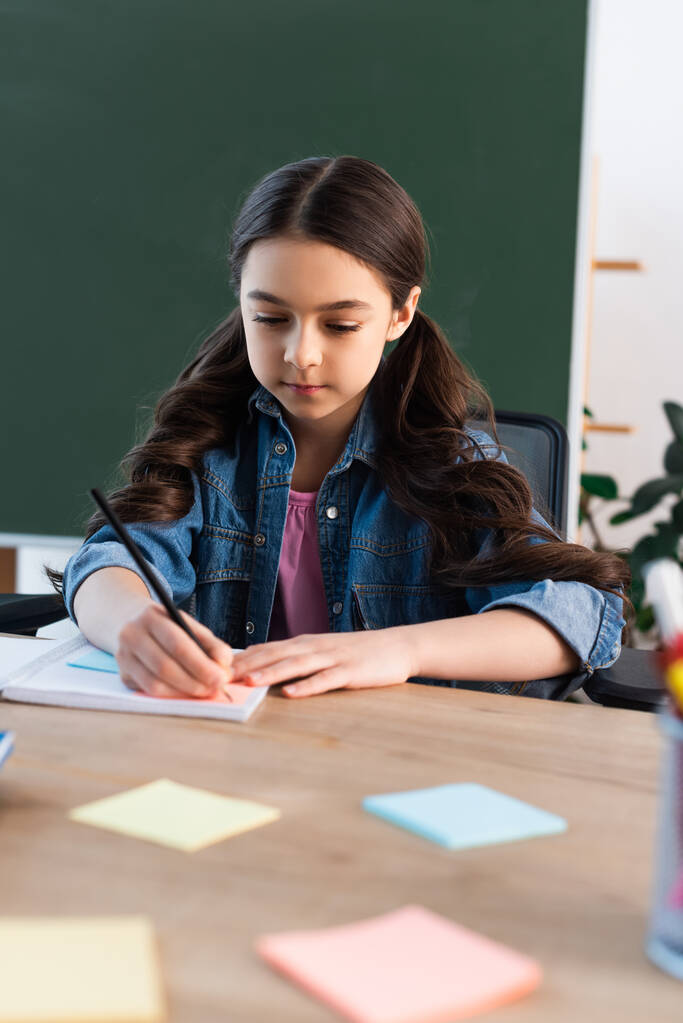 brunette meisje schrijven in notebook in de buurt wazig plakkerige notities en schoolbord - Foto, afbeelding