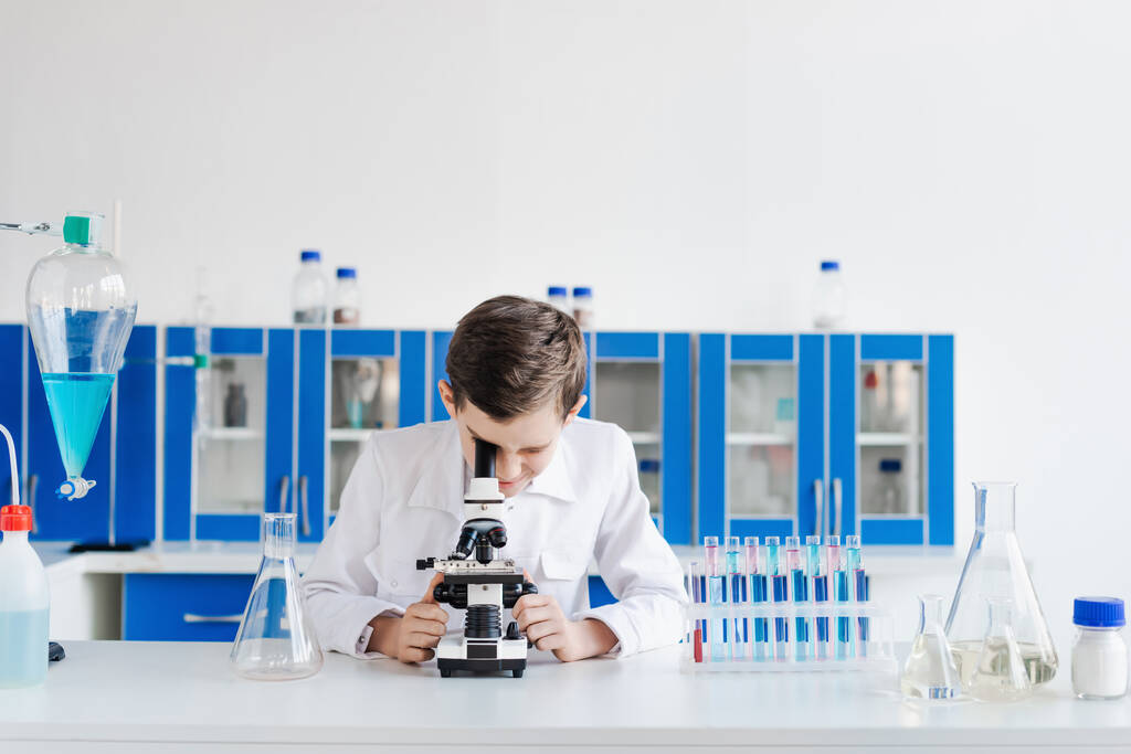 chlapec v bílém plášti při pohledu do mikroskopu v blízkosti zkumavek a baněk v laboratoři - Fotografie, Obrázek