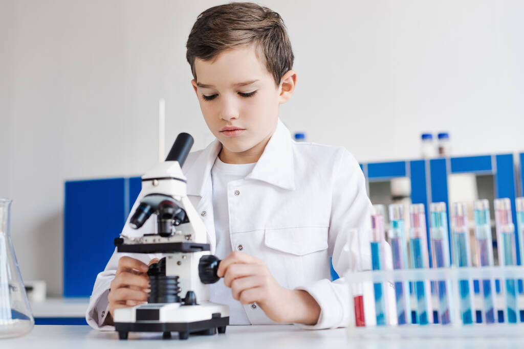 preteen αγόρι σε λευκό παλτό κοντά στο μικροσκόπιο και θολή δοκιμαστικούς σωλήνες στο εργαστήριο - Φωτογραφία, εικόνα