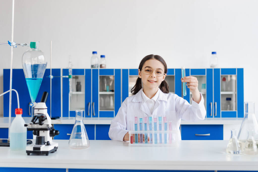 alegre chica en gafas y capa blanca sosteniendo tubo de prueba mientras está sentado cerca del microscopio y frascos - Foto, Imagen