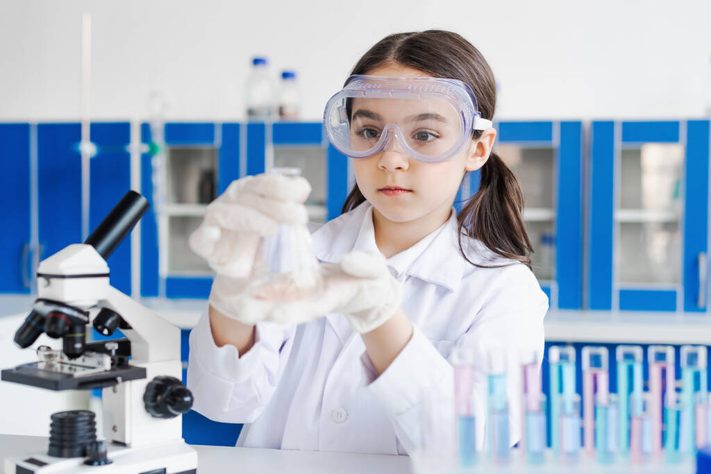 Девушка в очках держит фляжку возле микроскопа в лаборатории - Фото, изображение