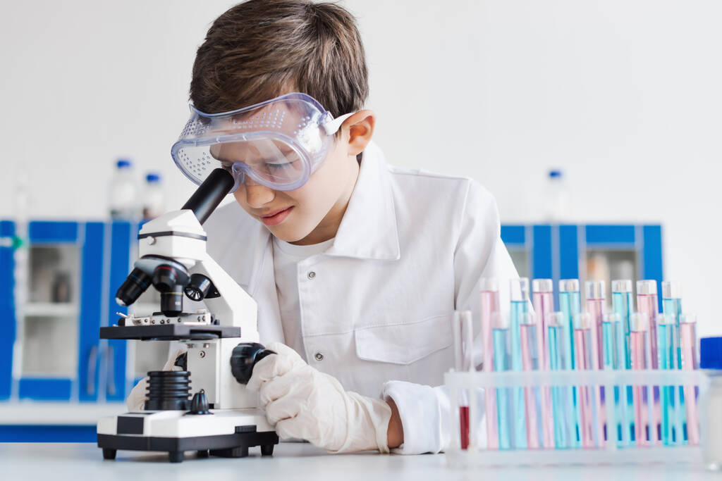 ребенок-подросток в очках и латексных перчатках смотрит в микроскоп рядом с пробирками  - Фото, изображение