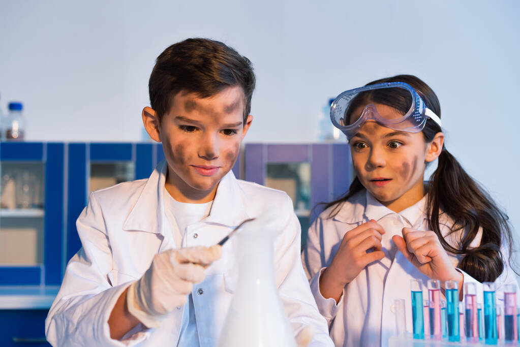 化学実験室で汚い顔をした男の子の手の中にぼやけたピンセットを見て怖い女の子 - 写真・画像