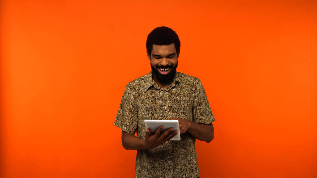 alegre joven afroamericano con barba sosteniendo tableta digital sobre fondo naranja  - Foto, Imagen