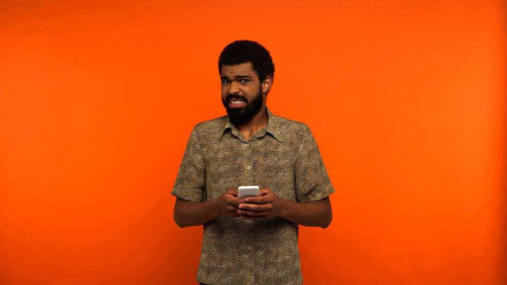 δυσαρεστήθηκε αφρικάνος Αμερικανός άνθρωπος μηνυμάτων smartphone σε πορτοκαλί φόντο  - Φωτογραφία, εικόνα