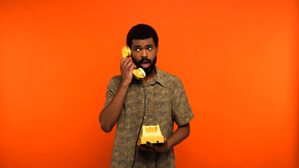 fiatal afro-amerikai férfi szakállas beszél sárga retro telefon narancs háttér  - Fotó, kép