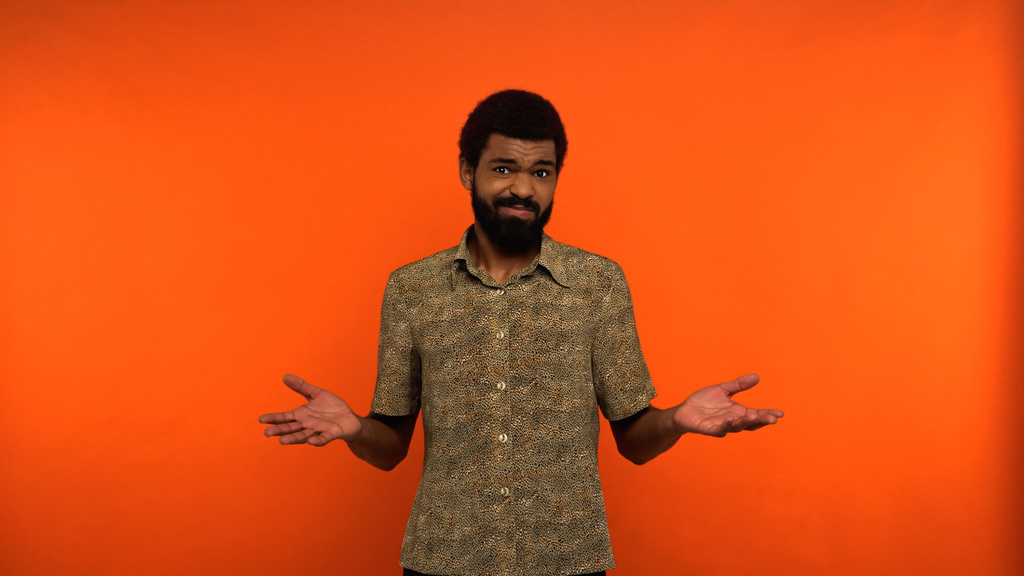 hombre afroamericano incierto en camisa mirando la cámara mientras gesticulaba sobre fondo naranja  - Foto, Imagen